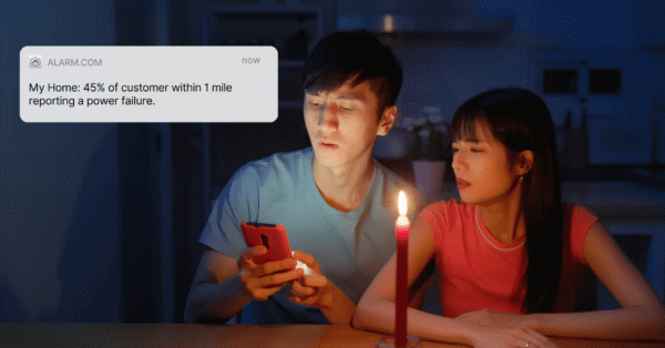 Couple sitting by candlelight during a power outage, looking at a phone notification from Alarm.com that reports a local power failure affecting 45% of customers.