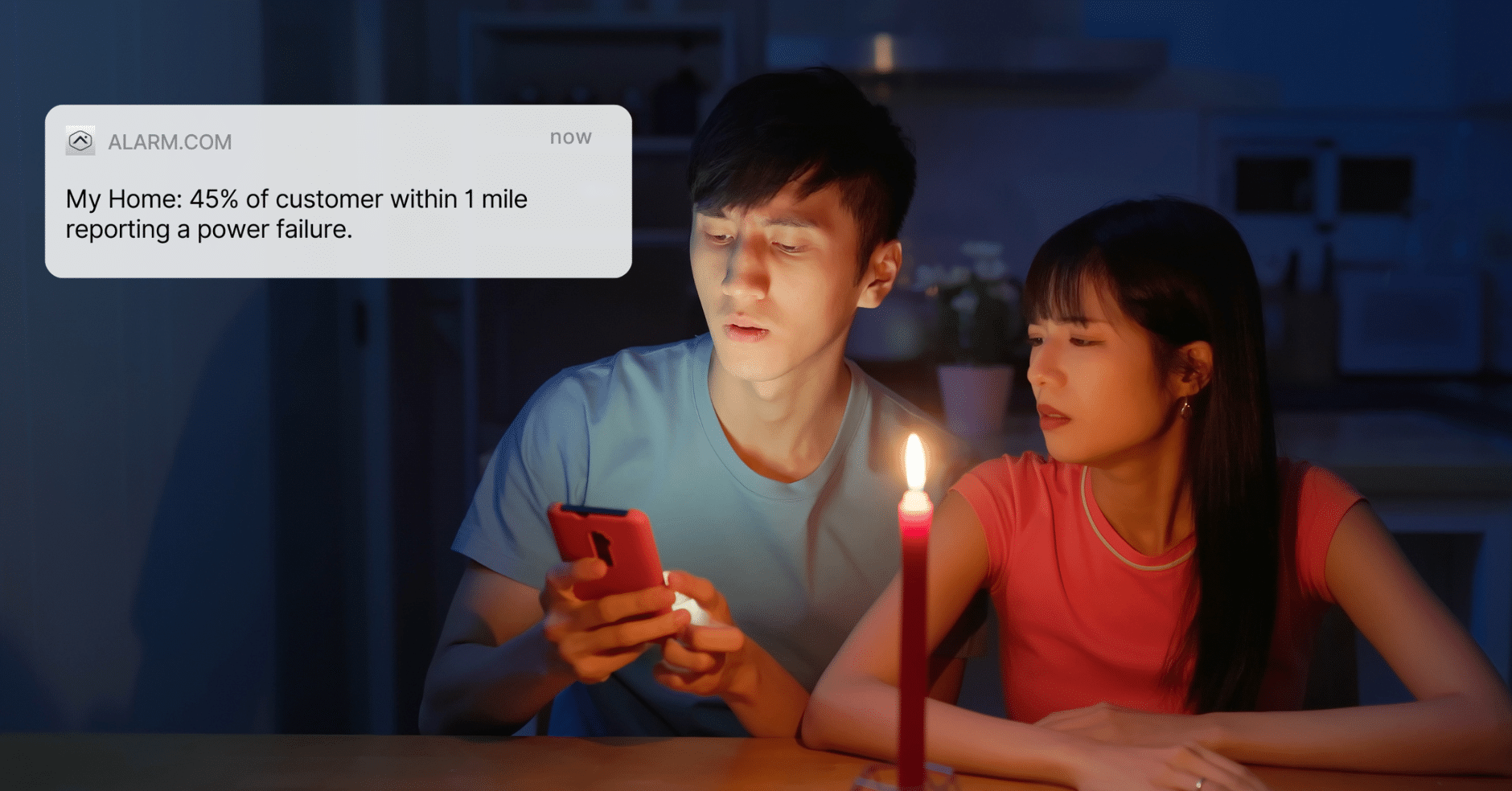 Couple sitting by candlelight during a power outage, looking at a phone notification from Alarm.com that reports a local power failure affecting 45% of customers.