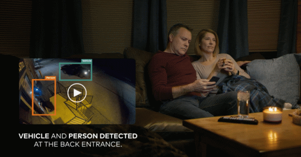 A couple sitting on a couch reviewing security camera footage and alerts on a mobile app, showing vehicle and person detection at a back entrance.
