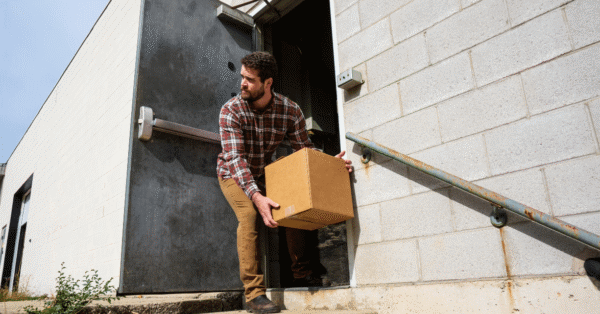 A man carrying a box exits a warehouse through a back door, representing employee theft and unauthorized inventory removal.