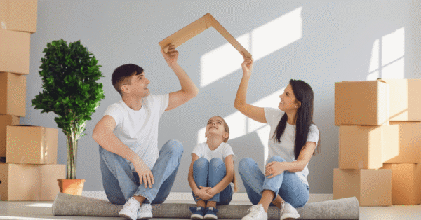 A young family sitting on the floor of their new home, holding a cardboard cutout of a roof over their heads, symbolizing safety and security.