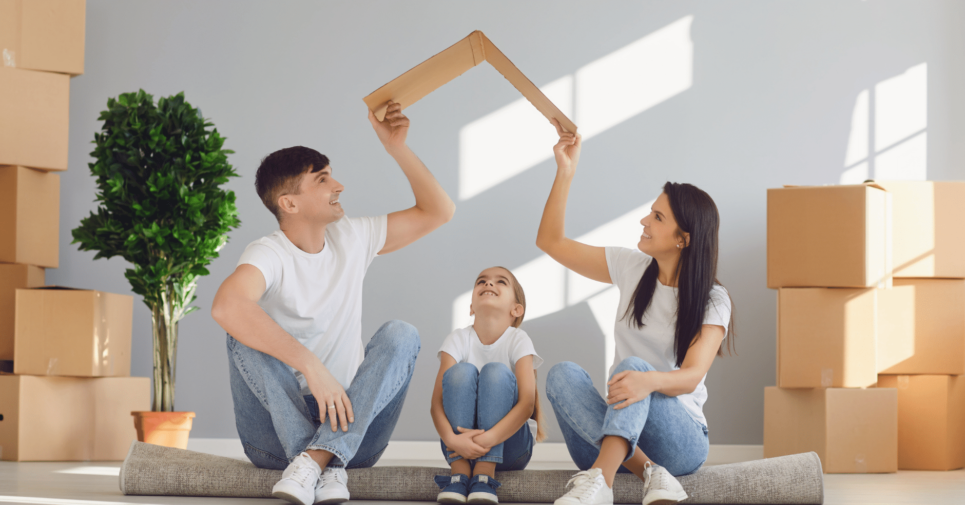 A young family sitting on the floor of their new home, holding a cardboard cutout of a roof over their heads, symbolizing safety and security.