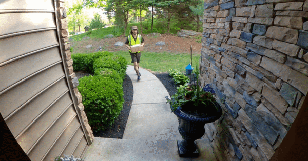 A delivery driver walking up a landscaped pathway toward a home, captured from the perspective of a video doorbell, highlighting its wide-angle surveillance capability.