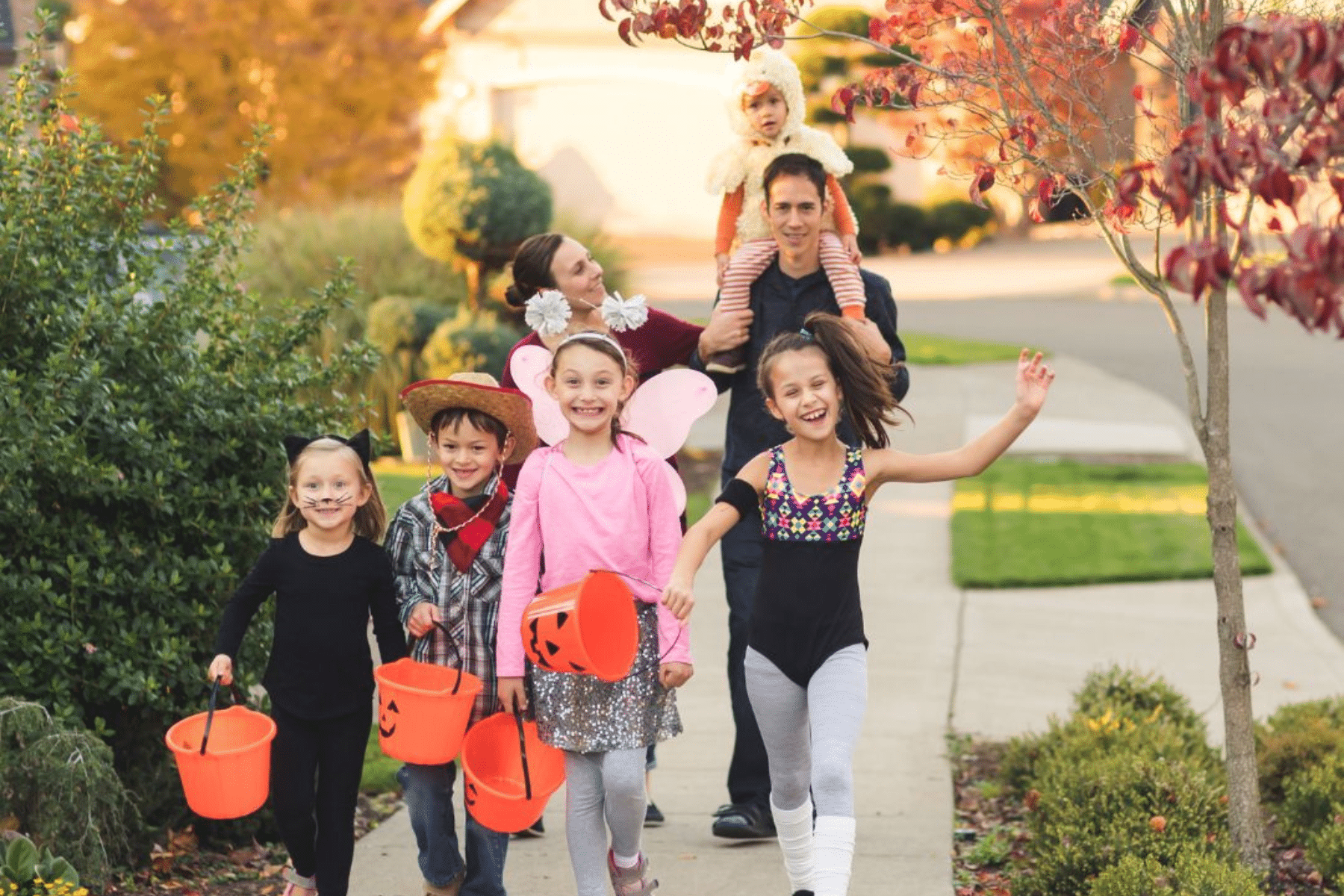 A group of children dressed in Halloween costumes, holding orange pumpkin buckets, walking joyfully down a neighborhood sidewalk with two parents and a younger child in a costume following behind.