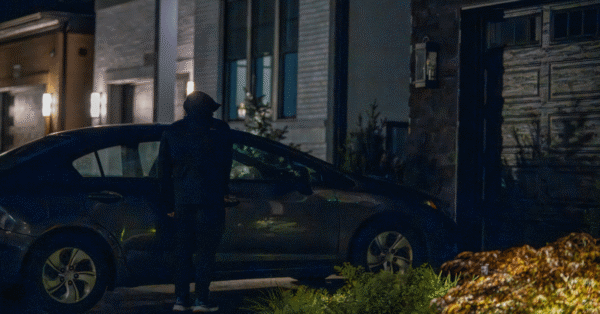A hooded figure attempting to break into a car parked outside a home at night, highlighting the importance of a reliable home security system to deter burglars.