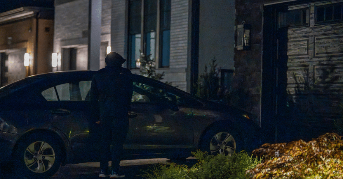 A hooded figure attempting to break into a car parked outside a home at night, highlighting the importance of a reliable home security system to deter burglars.