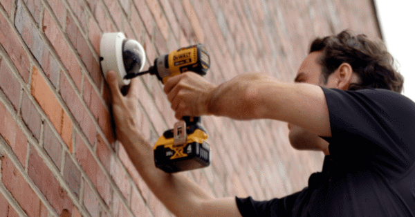 Professional technician installing a security camera on a brick wall with a drill.