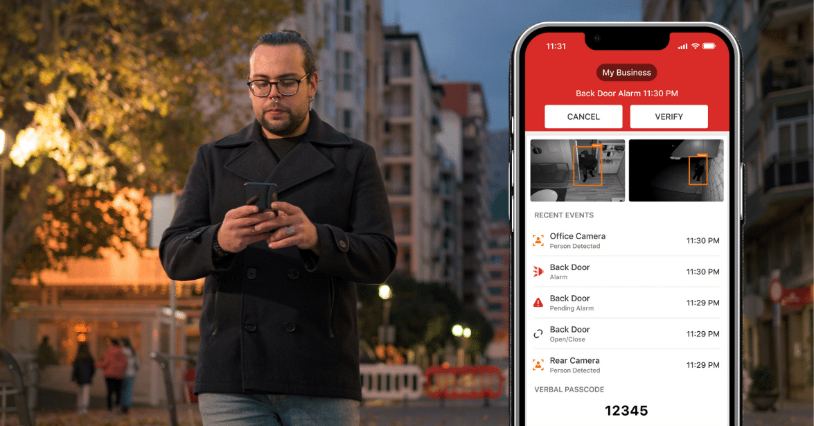 A man walking outdoors at night, checking his phone while a security app on the screen shows a back door alarm notification with options to 'Cancel' or 'Verify,' along with recent security events and camera footage.